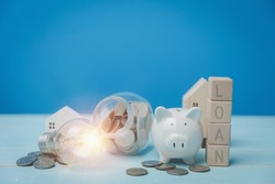 Text LOAN on wood blocks with a white piggy bank, and a light bulb. Behind have coins, a little wooden house on a blue desk. Financial, Investment estate, Loan, bank Concept.