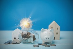 The light bulb on the bottom with coins, and a white piggy bank. Behind have a little wooden house on a blue desk. Financial, Investment estate, Loan, bank Concept.
