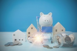 The light bulb with coins in the glass. Behind have a little wooden house, a white piggy bank on a blue shopping cart on a blue desk. Financial, Investment estate, Loan, bank Concept.