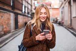 Young beautiful blonde and dreamy woman wearing casual terracotta coat writes sms online with smartphone chatting at street in autumn