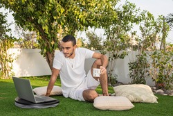 A young man in white clothes with a laptop is studying online sits in a home garden with coffee Work online from home Quarantine and self-isolation continues Coronavirus is still with us