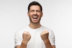 Closeup of emotional European man isolated on gray background showing white teeth while screaming with joy and victorious expression, holding hands in gesture of winner, looking happy
