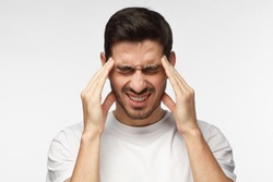 Portrait of young man isolated on grey background suffering from severe headache, pressing fingers to temples, closing eyes to relieve pain with helpless face expression