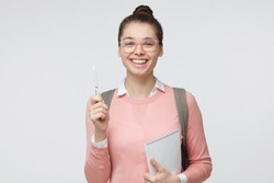 Studio shot of positive student girl isolated on grey background wearing glasses, pink sweater and backpack, pointing up with pencil, holding notebook, looking excited, full of energy and ideas