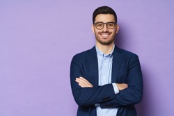 Young buisnessman wearing eyeglasses, jacket and shirt, holding arms crossed, looking at camera with happy confident smile, standing against purple background
