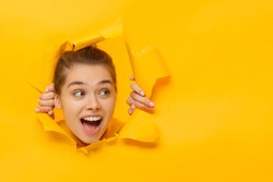 Young woman tearing paper and peeking out hole, curious about something on copy space on right, isolated on yellow background