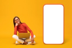Happy young woman sitting with laptop and looking at blank screen of huge phone, mockup for app, isolated on yellow background