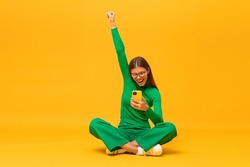 Winner. Excited happy business woman sitting on floor with phone, raising one hand in the air saying yes, isolated on yellow background