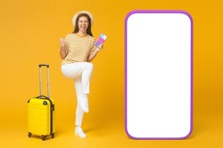 Happy excited girl in hat with suitcase heading for vacation holding cheap travel tickets, screaming yes, isolated on yellow, facing huge phone mockup with copy space screen for your content