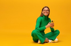 Dreamy student girl in stylish green clothes sitting with crossed legs on floor using smartphone, having rest on yellow copy space background holding phone in hands, looking up aside