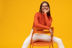 Studio shot of charismatic attractive stylish millennial female student with pensive dreamy face sitting on chair leaning on chairback, isolated on yellow background, looking aside smiling