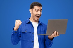 We got a winner! Excited young man in blue shirt looking at laptop screen with WOW expression, isolated on blue background