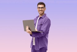 Young man standing holding laptop and looking at camera with happy smile, isolated on purple background