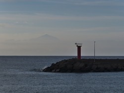 View of a beacon at port of La Aldea de San Nicolas on the western coast of Gran Canaria, Spain with the silhouette of Tenerifa island with mountain Pico del Teide in background on misty day.