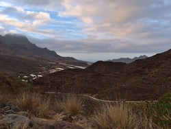 Landscape of the western coast of island Gran Canaria, Spain with remote village La Aldea de San Nicolas surrounded by rugged mountains on cloudy day with road dry plants in front.