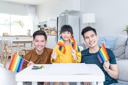 Portrait of Asian LGBTQ men gay family with daughter in living room. Attractive handsome male couple take care and spend time with little cute child holding LGBTQ rainbow flag and looking at camera.