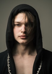 Handsome boy in black hood with medium length layered dark hair with long chain around his neck standing isolated on gray background, youth fashion concept.