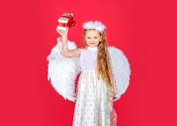 Child angel little girl with present gift, studio portrait. Little angel, happy fairy with white wings holds gift. Cute angel child girl with angels wings, isolated on red. Valentines day.