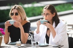 Two beautiful girls with cups coffee in summer cafe. Girl friends in cafe outdoor. Outdoors portrait of two young beautiful women friends drinking coffee.
