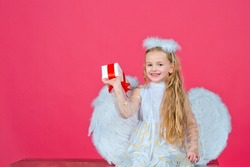 Angel kids, happy fairy with present gift, studio portrait. Little blonde angel with white wings holds gift. Cute angel child girl with angels wings, isolated on red. Valentines day.
