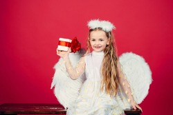 Kid girl angel with present gift, studio portrait. Little angel with white wings holds gift, happy fairy. Cute angel child girl with angels wings, isolated on red. Valentines day.