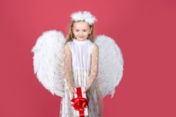 Kid girl angel with present gift, studio portrait. Little angel with white wings holds gift. Cute angel child girl with angels wings, isolated on red. Valentine gift card.