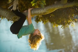 Childhood leisure, happy kids climbing up tree and having fun in summer park. Young boy playing and climbing a tree and hanging upside down. Teen boy playing in a park.