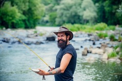 Hipster bearded man catching trout fish. Young man fishing. Fisherman with rod, spinning reel on river bank. Man catching fish, pulling rod while fishing on lake. Wild nature.