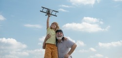 Grandson child and grandfather with toy plane over blue sky and clouds background. Two men generation grandfather and grandson playing outdoors. Generational family.