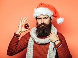 young handsome bearded santa claus man with long beard in red sweater and new year hat in scarf holds decorative christmas or xmas ball on orange studio wall background