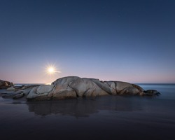 A long exposure of the Bloodmoon rising over rocks on a beach during the lunar eclipse at blue hour.  The moon cast an incredible shadow of the rock creating a reflection underneath the stars