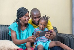 
A three member family curdled and sitting together on a sofa with a mobile smartphone in their hands, the group consist of a little African girl, woman and a man 