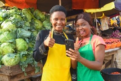 Two happy African business women or female traders wearing colorful aprons while standing at a vegetable stall in a market place with a smart phone with them