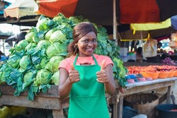 Happy African business woman or female trader wearing a green apron, doing thumbs up gestures while standing at her stall of vegetables in a market place