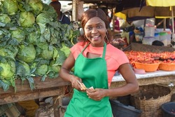 Happy African business woman or female trader wearing a green apron and standing at her vegetable stall in a market