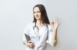 young female doctor in doctor's uniform and stethoscope, smiling, makes a positive sign with hand and fingers. showing the OK sign, looking at camera and smiling