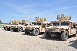 Desert tan US Military armored  High Mobility Multi-Wheeled Vehicles (HMMWV) often used in the wars in Afghanistan and Iraq parked in a gravel lot with a blue sky and green grass in the background.