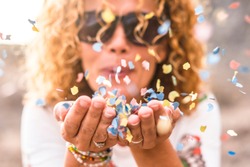 Close up of woman hands and beautiful lady blowing out coloured carnival party confetti - focus on colors papers and happiness and joyful lifestyle concept for happy people outdoor