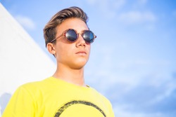 Handsome young boy man with blonde hair and black sunglasses stand and pose with yellow shirt and blue sky background - caucasian guy summer portrait