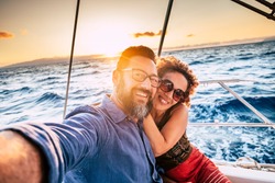 Happy and cheerful people enjoying the travel and trip on a sail boat with ocean and sunset sunlight in background - traveler lifestyle for adult man and woman smiling doing a selfie picture