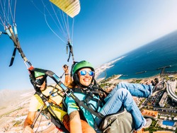 Cheerful happy woman to paraglyde experience with pilot - couple having fun in the air paraglyding over the city with coastline view - tourist and summer holiday vacation active people