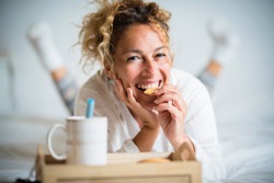 Portrait of adult beautiful woman eating cookie in morning breakfast in the bedroom - home or hotel wake up day with pretty female people lay down with biscuit and tea - cheerful and happy face lady