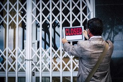 Rear view of businessman putting closed sign on display on locked shop or office due to outbreak of coronavirus pandemic. Government shutdown of non essential services. Closing due to bankruptcy