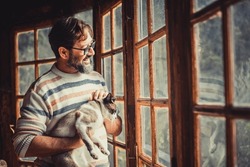 Adult happy mature man with a cat look outside the windows at home enjoying indoor leisure relax and activity alone. Hipster male people with window light smiling in living room