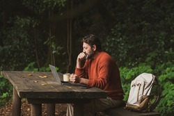Mature worker man sit down on a wooden bench and work on the table in the middle of park outdoors forest woods alone. Backpack and laptop computer with people busy in modern job connection activity