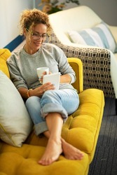 Pretty woman enjoy relax at home reading a book on tabler reader and drinking a tea or coffe sitting on a yellow sofa in living room - modern online library store and female people using device gadget
