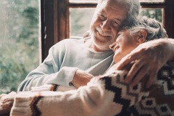 Old senior couple in love hug and embrace with romance together at home with outside view in windows background - happy mature retired people lifestyle enjoying caring each other and smiling