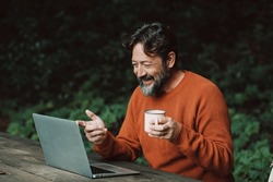 Adult hipster bearded man enjoying video call conference outdoors in the nature using laptop computer. Concept of modern people and digital nomad smart working job activity outdoors