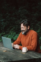 Happy free adult man work outdoors on a table with nature in background - concept of digital nomad modern online lifestyle people - mature male with beard use laptop computer in the woods smiling 