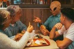 Happy family with ufo alien estraterrestrial guest enjoy together hamburgers in fast food. Concept of friendship and diverse people having fun together. Alien explaining at humans